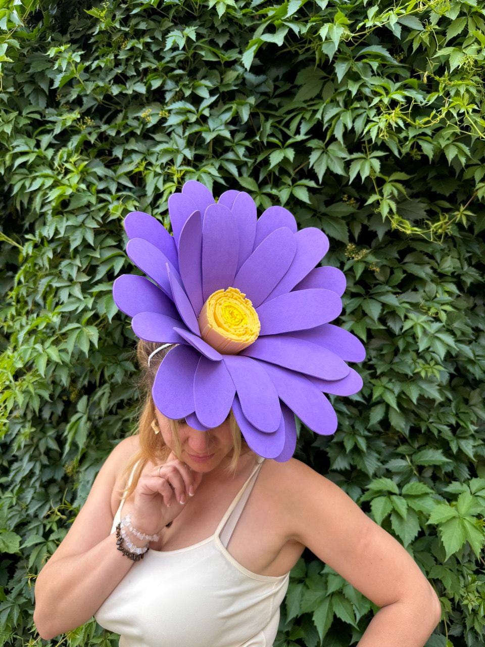 May include: A large, purple flower hat with a yellow center. The hat is made of layered petals and is worn by a person. The background is a wall of green leaves. The hat is a unique accessory.