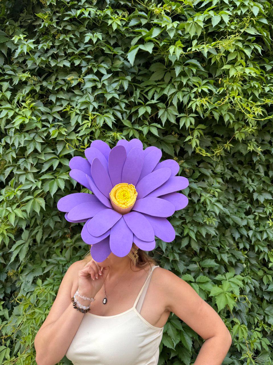 May include: A large, purple paper flower with a yellow center covers the face of a person wearing a white tank top. The flower has multiple layers of petals. The background is a wall of green leaves.