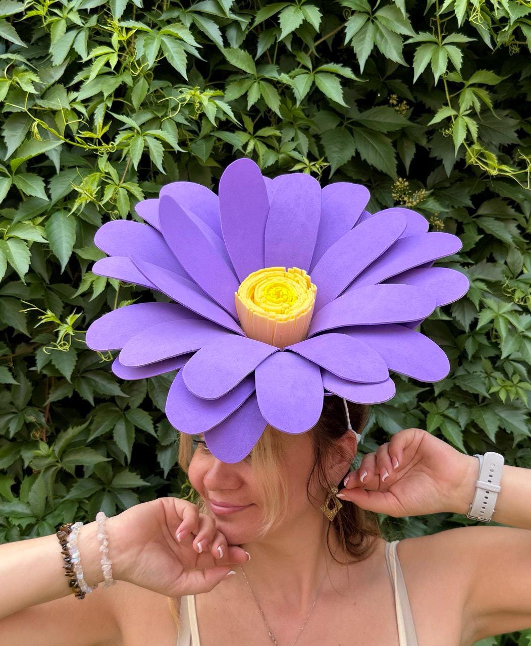 May include: A large, purple flower headpiece with a yellow center. The petals are layered and the headpiece is worn on the head. The background is a lush green wall of leaves.