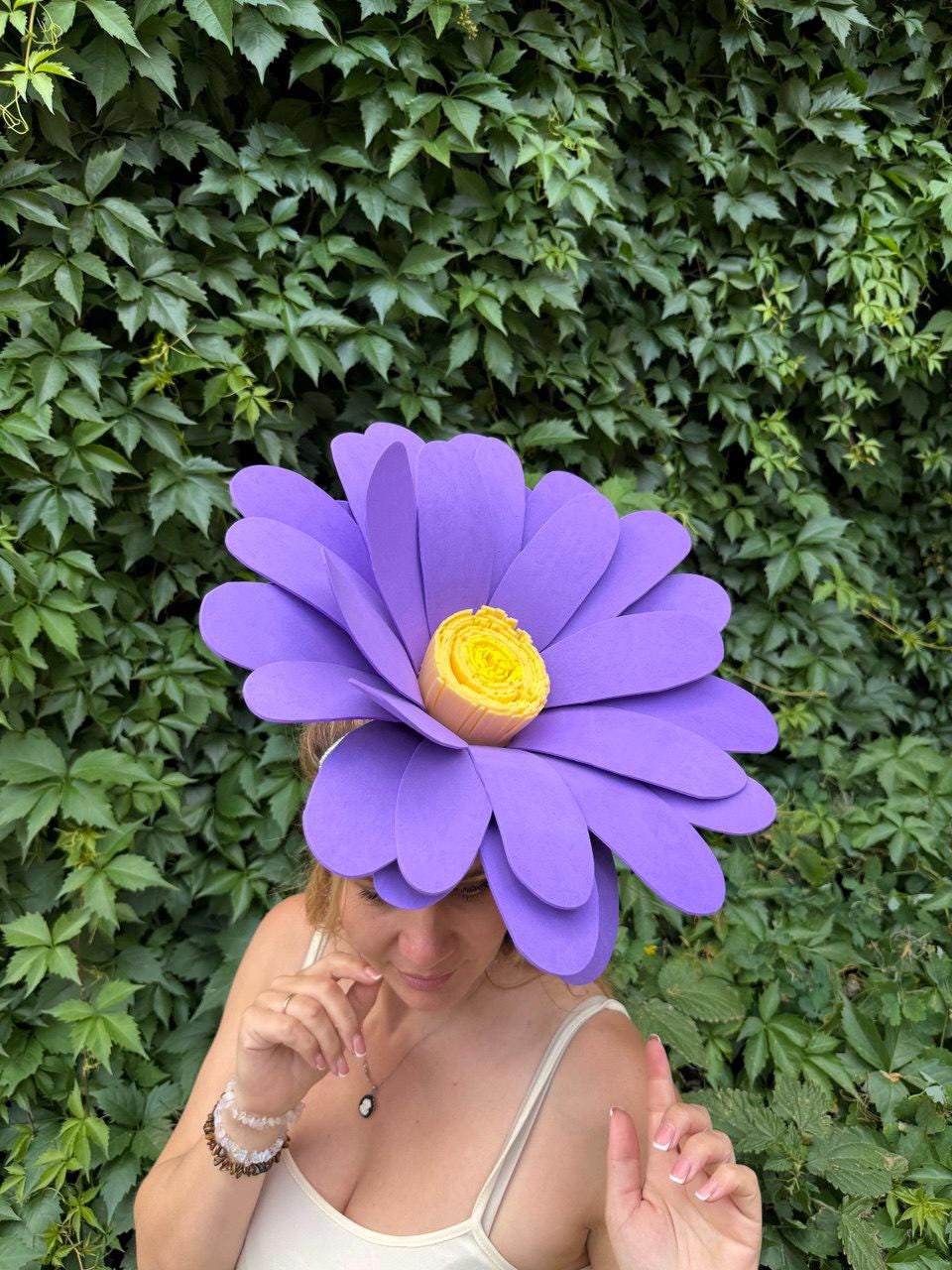 May include: A large, purple flower-shaped hat with a yellow center. The hat is made of layered petals and is worn on the head. The background is a wall of green leaves.