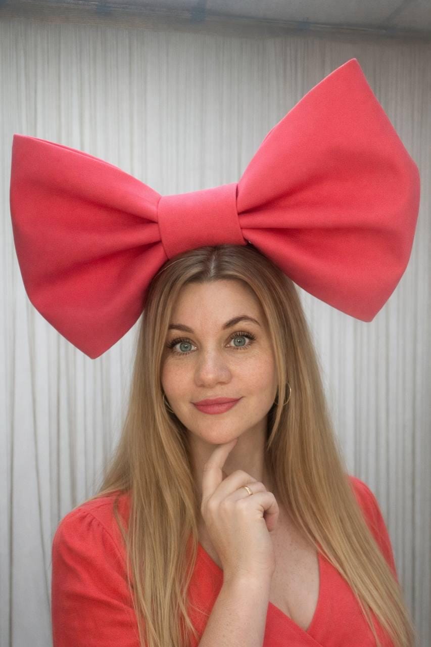 May include: A large, coral-colored bow headband. The bow is the focal point, positioned atop the head. The person in the image is wearing a coral-colored top. The background is a white, textured curtain.