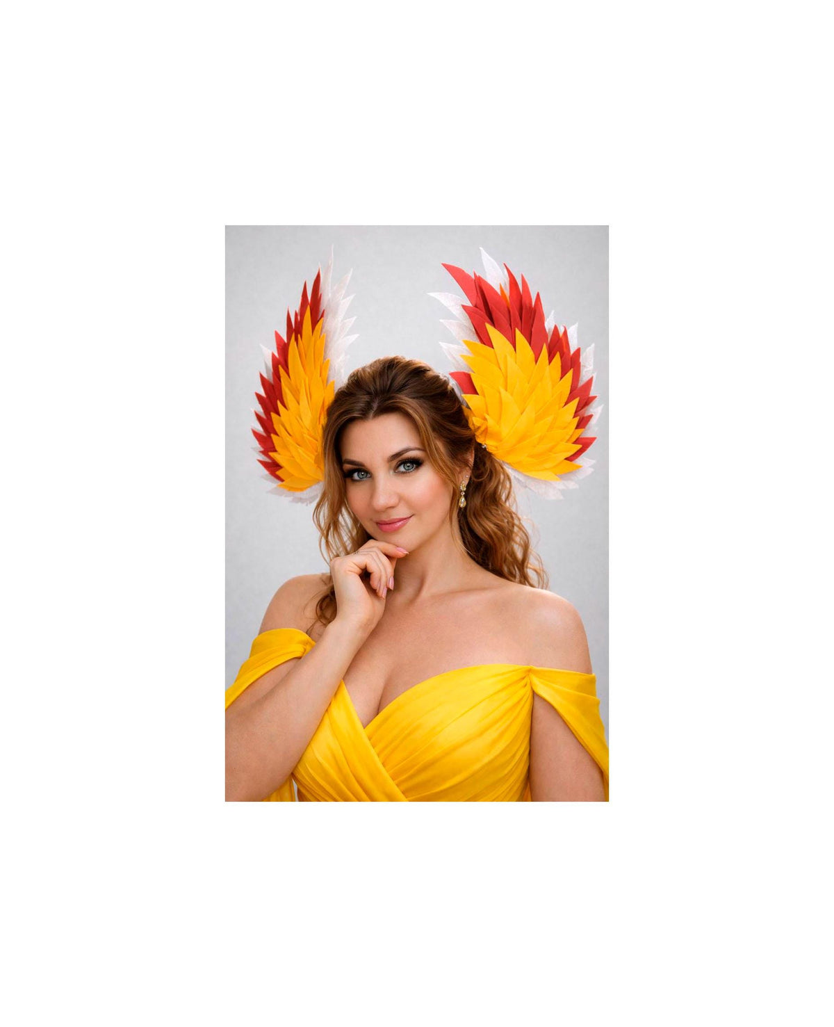 May include: A woman wearing a yellow off-the-shoulder dress and a pair of colorful, feathered wing-shaped headpieces. The headpieces feature red, yellow, and white feathers. The woman is smiling and looking at the camera.