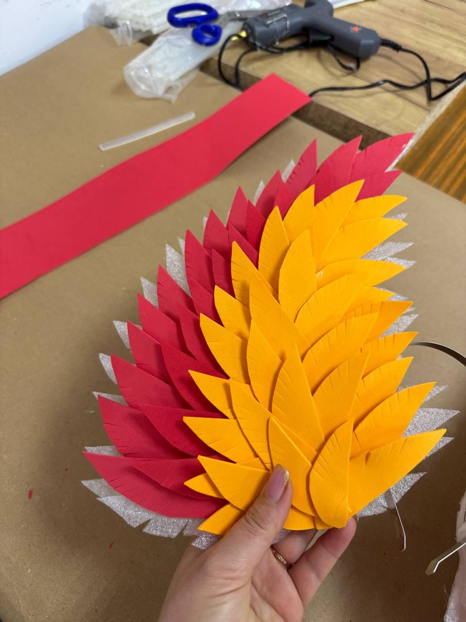 May include: A handmade, layered feather-shaped craft piece. The design features alternating rows of red and yellow foam leaves, creating a vibrant, textured effect. The craft is held by a hand, with craft supplies visible in the background.