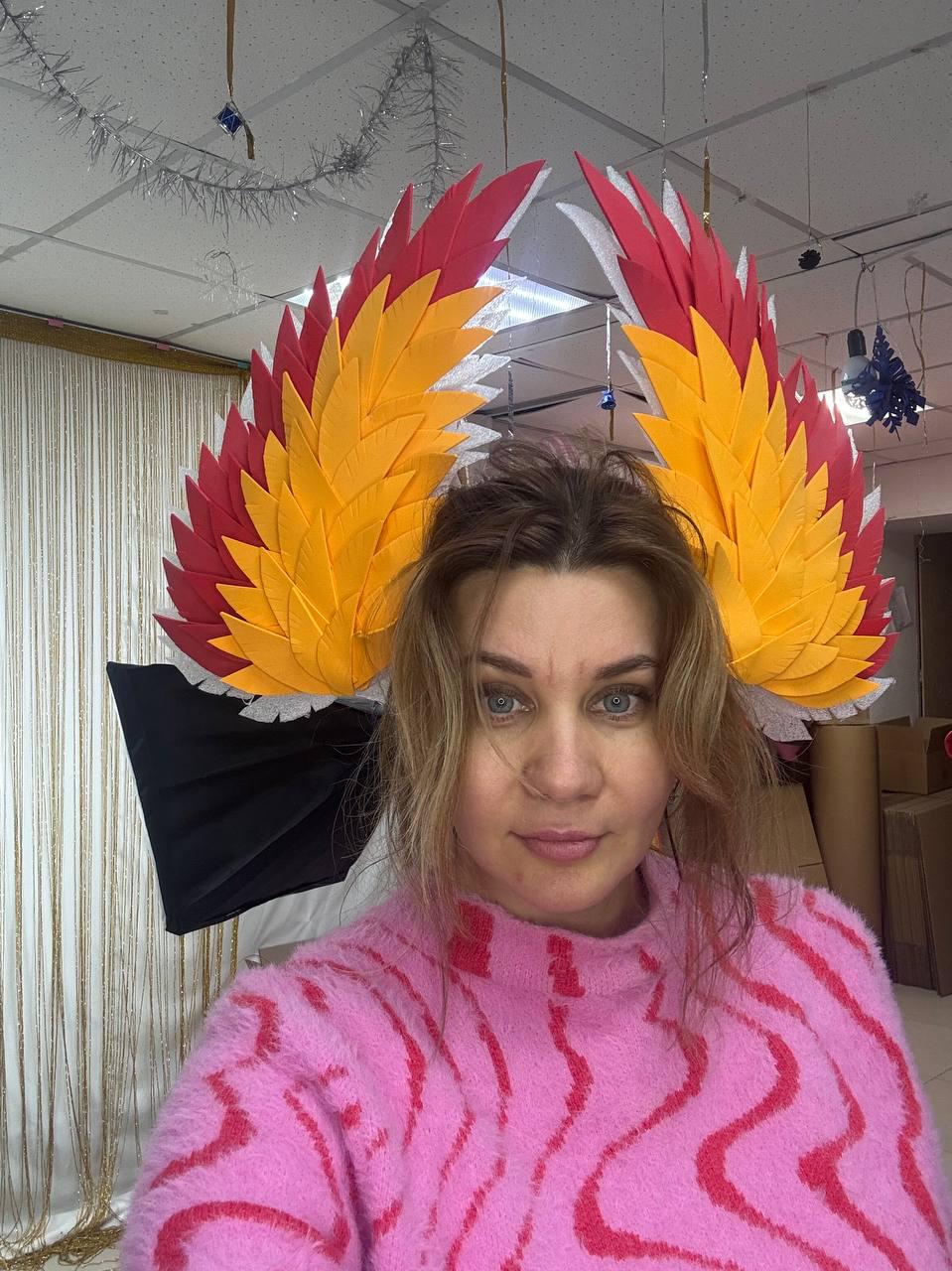 May include: A person wearing a pair of large, colorful, handmade wings. The wings feature red, yellow, and white feathers. The person is wearing a pink sweater with a red wavy pattern.