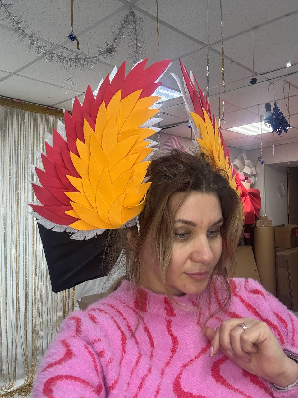 May include: A vibrant headpiece with layered felt feathers in red, yellow, and white, resembling wings. The headpiece is attached to a black base. The person in the image is wearing a pink sweater.
