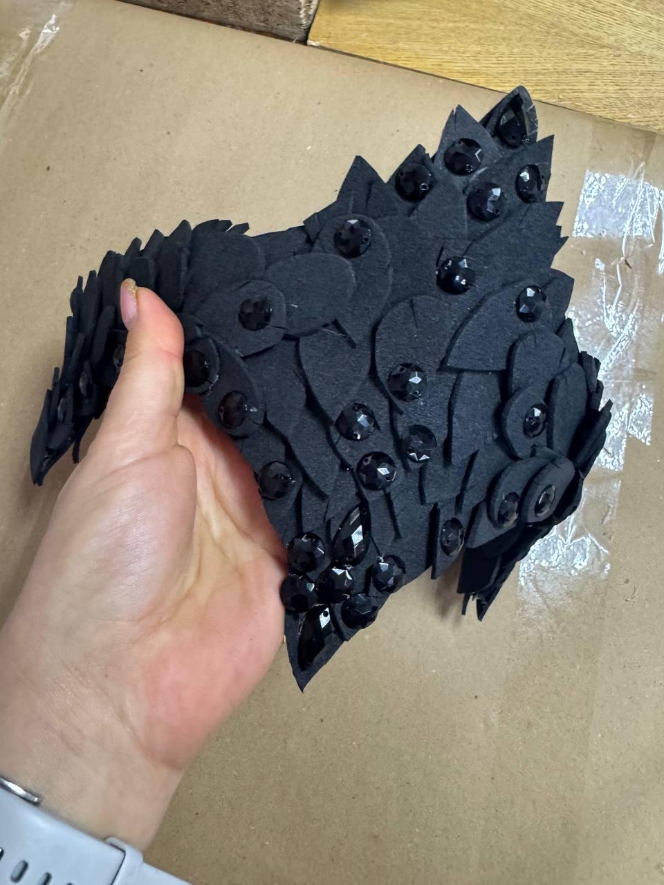 May include: A black, decorative headpiece with a layered, petal-like design. The headpiece is adorned with numerous black, faceted gemstones. The item is held in a person's hand.
