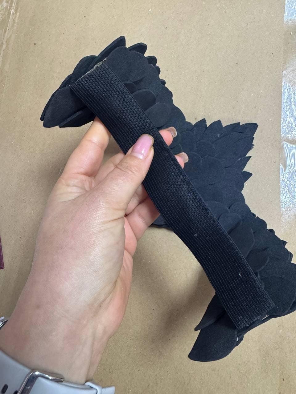 May include: A black, feather-like accessory with an elastic band. The accessory is held in a hand, and the feathers are layered to create a textured appearance. The background is a neutral tan color.