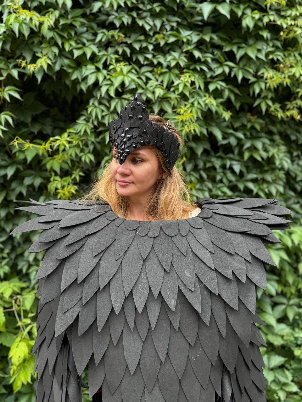 May include: A black costume featuring a feathered shoulder piece and a matching crown. The crown is adorned with small, dark embellishments. The costume is set against a backdrop of green foliage, creating a striking contrast.