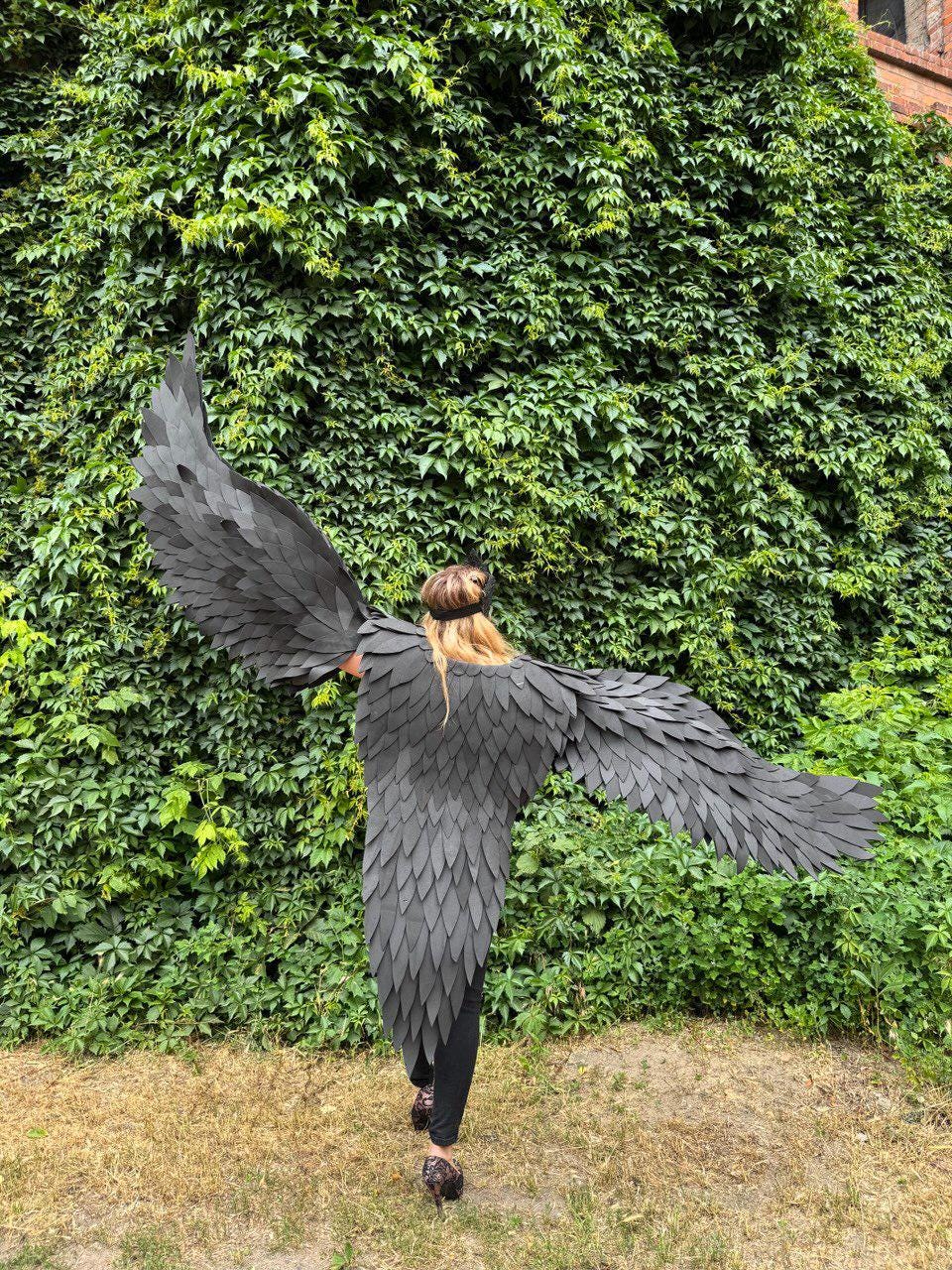 May include: A person wearing large, black, feathered wings, standing in front of a wall of green foliage. The wings are detailed with overlapping, textured segments, creating a dramatic effect. The wings extend outwards, showcasing their full span.