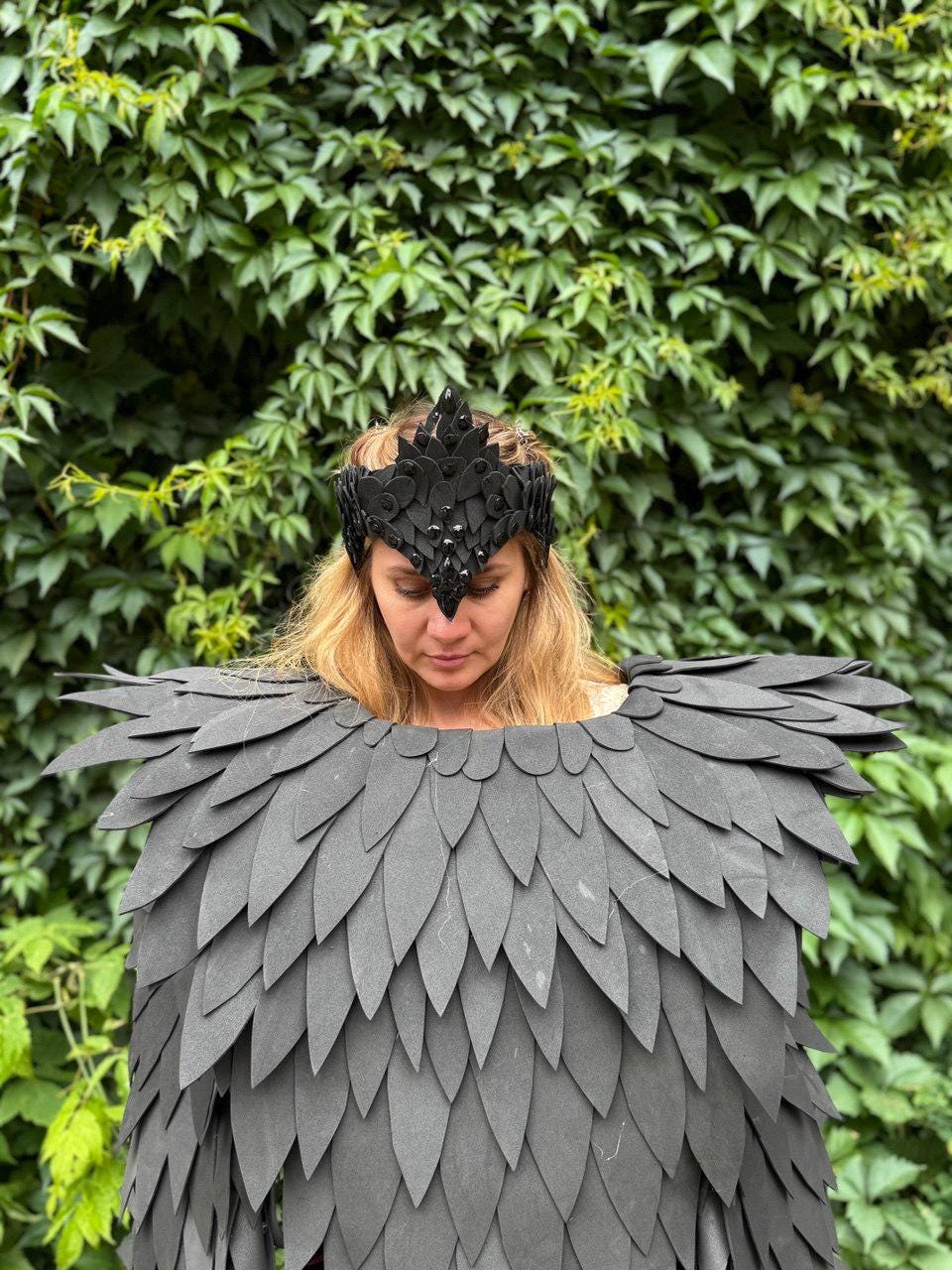 May include: A black crown and matching shoulder piece with a layered feather design. The crown has a pointed center and is adorned with small, dark embellishments. The shoulder piece is made of overlapping, dark gray, leaf-shaped pieces.