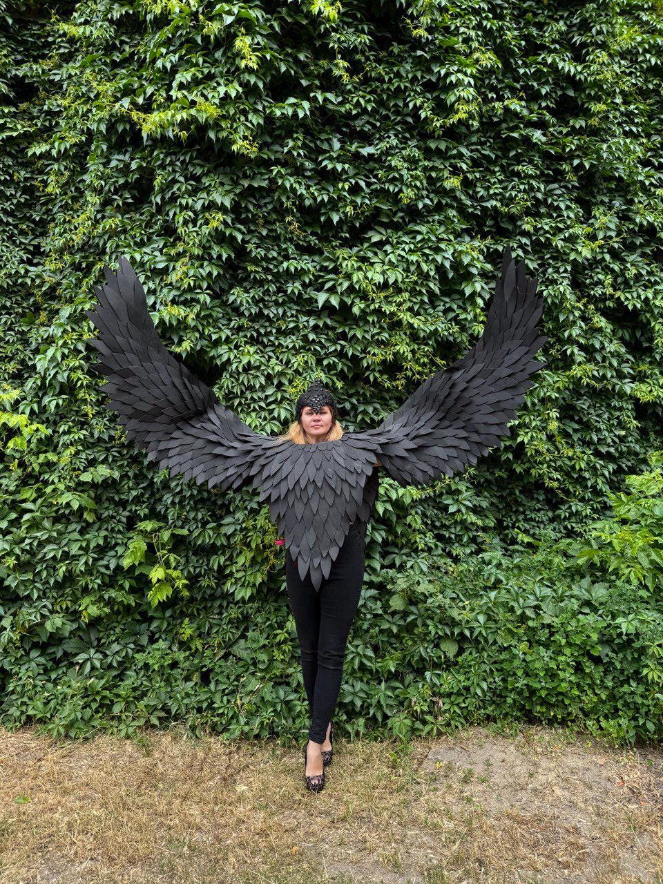 May include: A person wearing a black costume with large, outstretched wings. The wings and the upper body piece are made of layered, textured material. The person is standing in front of a wall of green foliage, wearing black pants and a black headpiece.