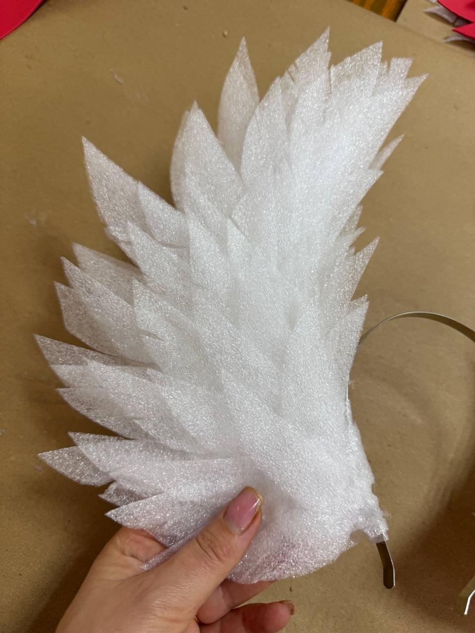 May include: A white, feather-shaped craft element made of textured foam. The foam is cut into individual feather shapes and attached to a thin, curved metal band. The item is held by a hand, suggesting it is a costume or decorative piece.