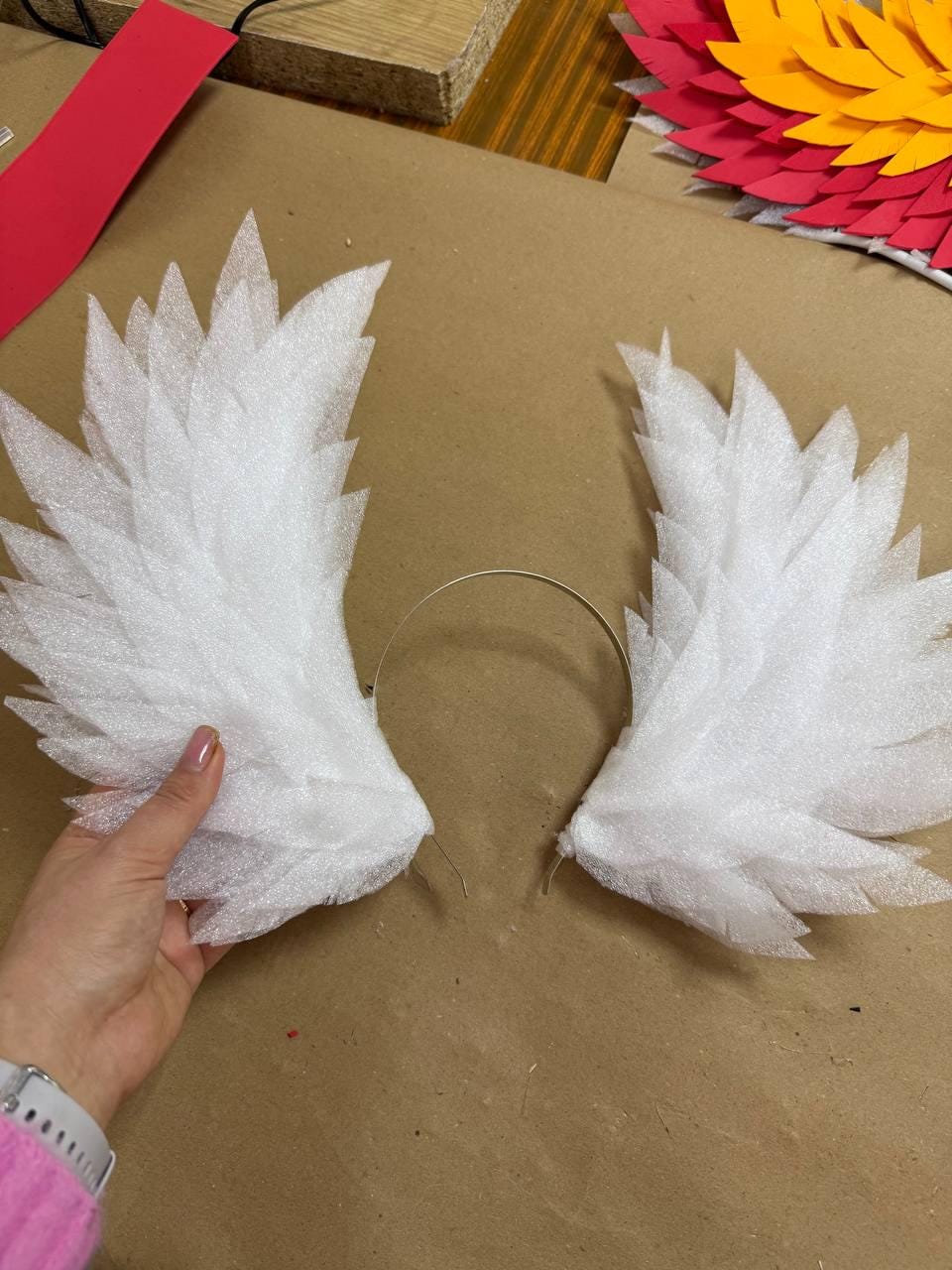 May include: White foam angel wings attached to a thin, silver-colored headband. The wings are made of layered, pointed foam pieces, creating a feathered effect. The wings are displayed on a brown surface.
