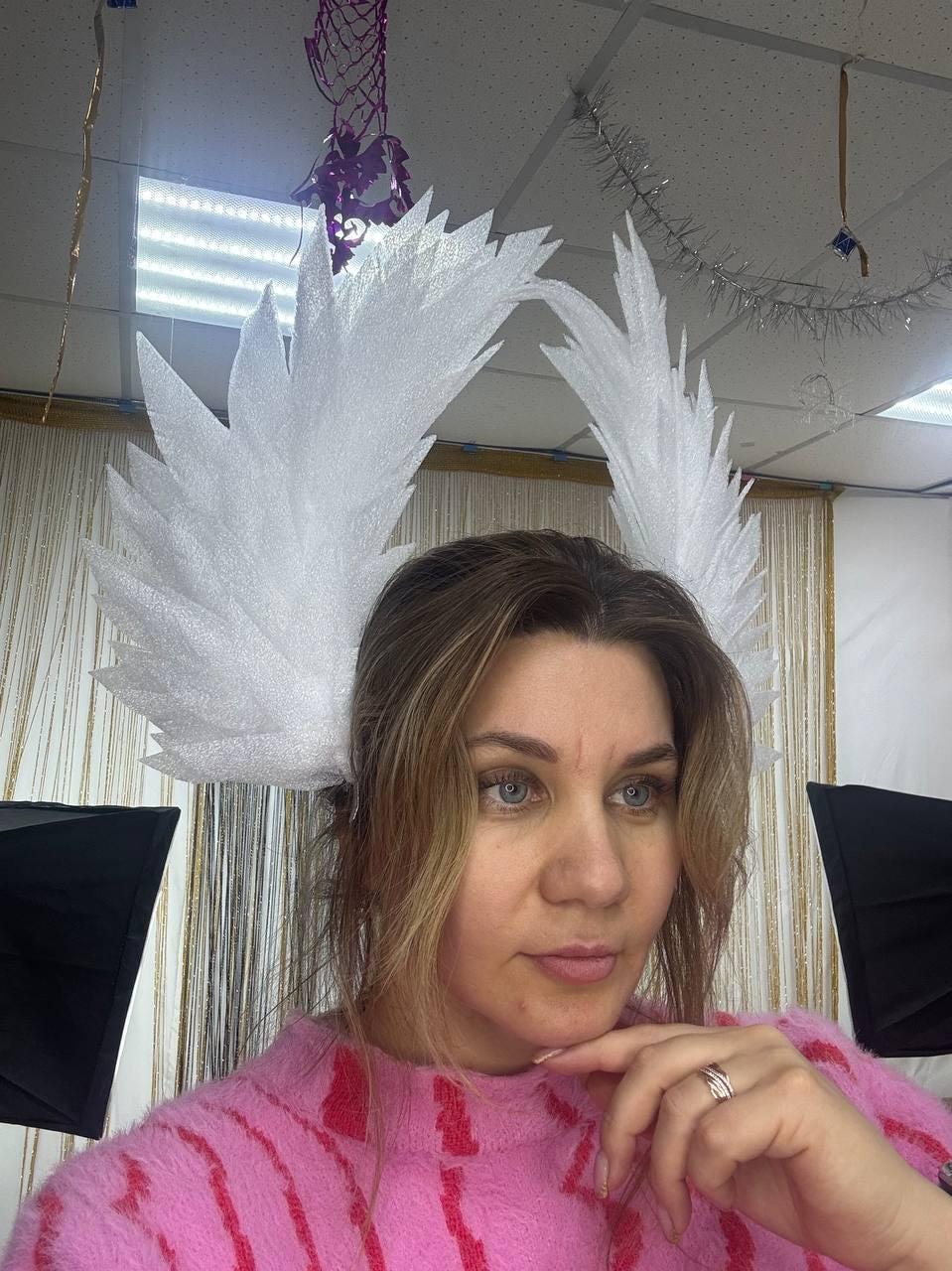 May include: A white, feathery angel wing headpiece. The headpiece is worn by a person wearing a pink sweater. The wings are large and textured, with a pointed, layered design. The background includes studio lighting and decorative elements.