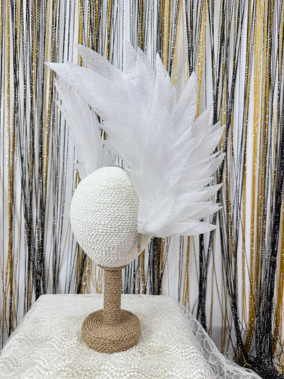 May include: A white, decorative headpiece with a textured, oval base and a feathery, fan-shaped accent. The base is mounted on a brown, rope-wrapped stand. The background features black, gold, and white vertical strands.