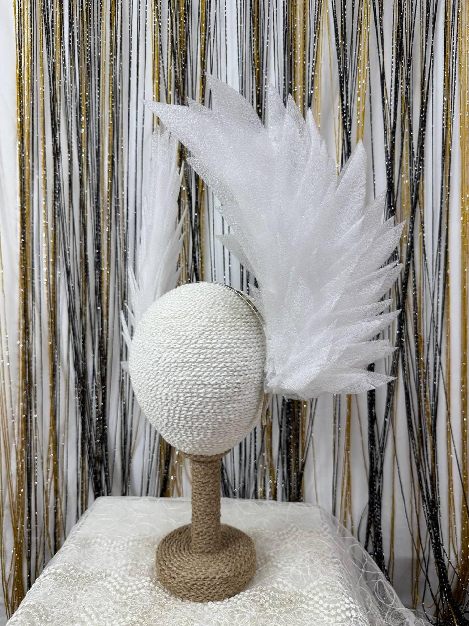May include: A white, textured head form with a feather-like wing design. The head form is mounted on a jute-wrapped stand, set against a backdrop of gold, black, and white decorative strands. The overall theme is a decorative headpiece.