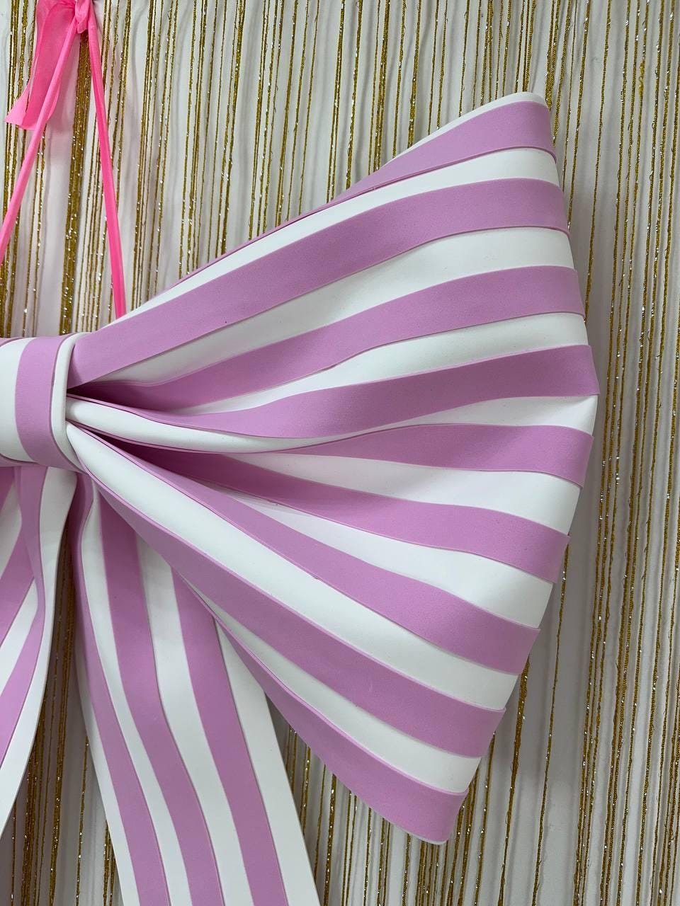 May include: A large, decorative bow with alternating pink and white stripes. The bow is tied in the center and has long, flowing tails. A pink ribbon is attached to the top for hanging. The background is a gold and white curtain.
