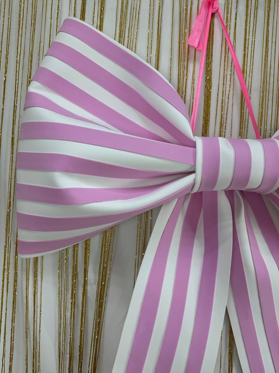 May include: A large, decorative bow with alternating pink and white vertical stripes. The bow is made of a smooth material and is suspended by a bright pink ribbon. The background features a white curtain with gold strands.