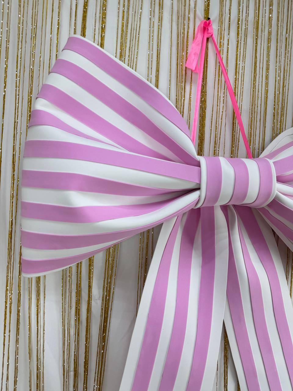 May include: A large, decorative bow with alternating pink and white stripes. The bow is made of a smooth material and has a bright pink ribbon for hanging. The background is a white curtain with gold vertical strands.