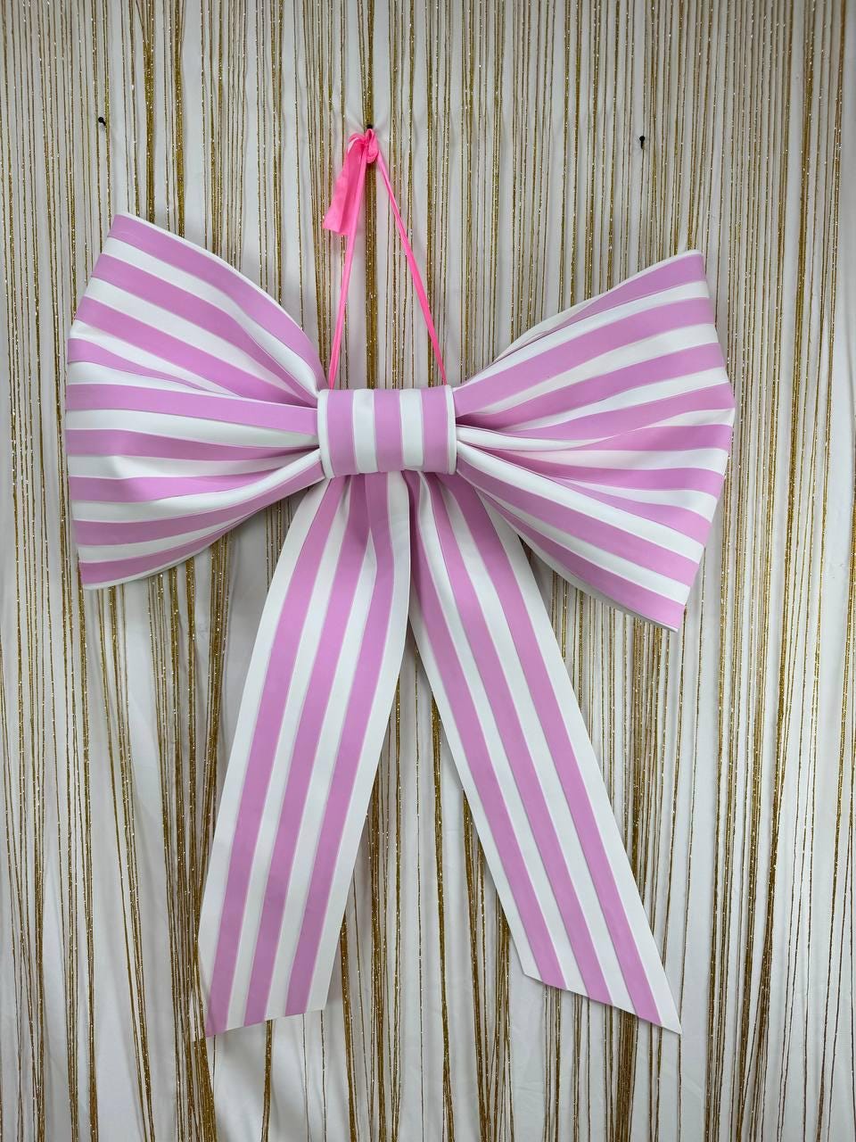 May include: A large, decorative bow with pink and white stripes. The bow is suspended by a pink ribbon. The bow is a festive decoration, suitable for parties, gifts, or home decor.