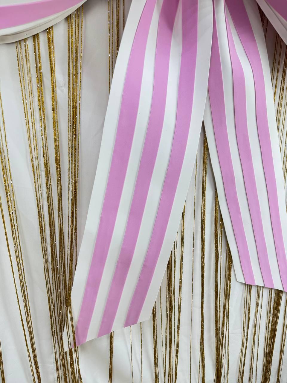 May include: A decorative backdrop featuring a white fabric with vertical gold glitter strands. A large bow with pink and white vertical stripes is draped over the backdrop, creating a festive and elegant look.