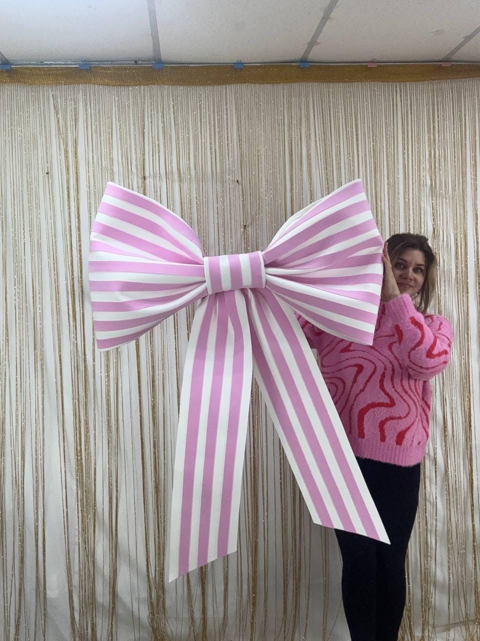 May include: A large, decorative bow with pink and white stripes. The bow is oversized and has long, flowing tails. The bow is a festive decoration, suitable for parties, events, or as a photo prop. The bow is made of a sturdy material.
