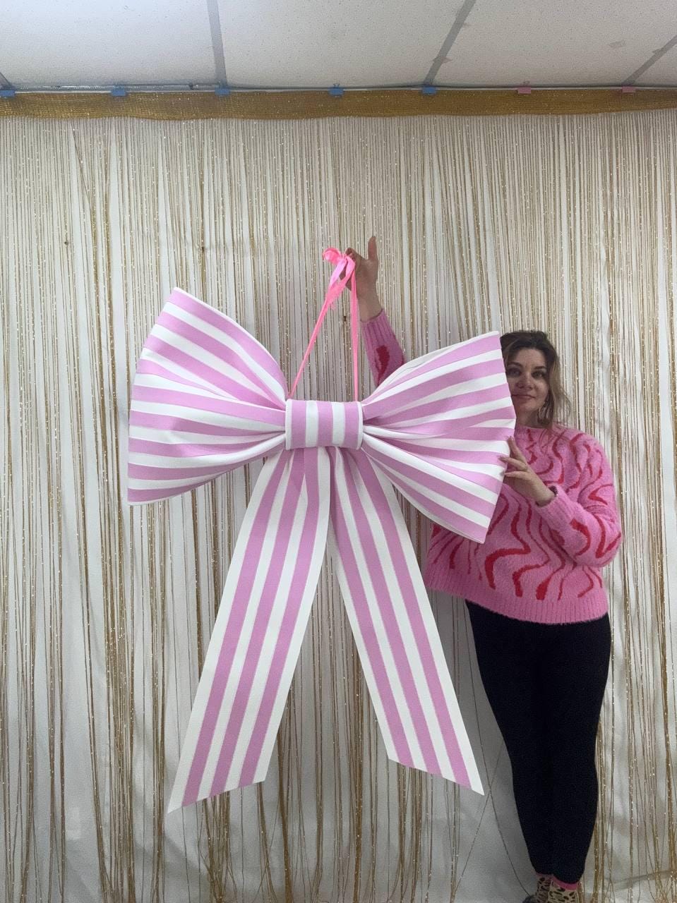 May include: A large, decorative bow with pink and white stripes. The bow is suspended by a pink ribbon. The bow is a festive decoration, suitable for parties, events, or as a unique gift wrapping accent.