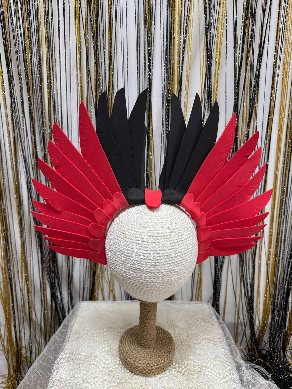 May include: A vibrant headdress featuring red and black feathers. The feathers are arranged in a wing-like design, attached to a black headband. The headdress is displayed on a white, spherical form.