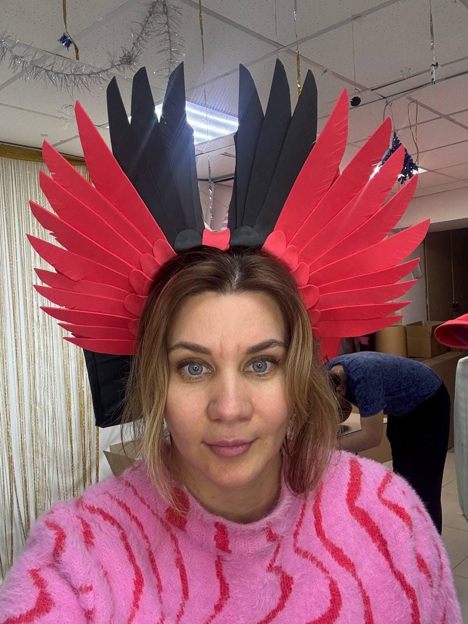 May include: A striking headpiece with large, stylized red and black feather-like elements. The headpiece is worn by a person wearing a pink sweater. The design suggests a theatrical or costume accessory, possibly for a festival or performance.