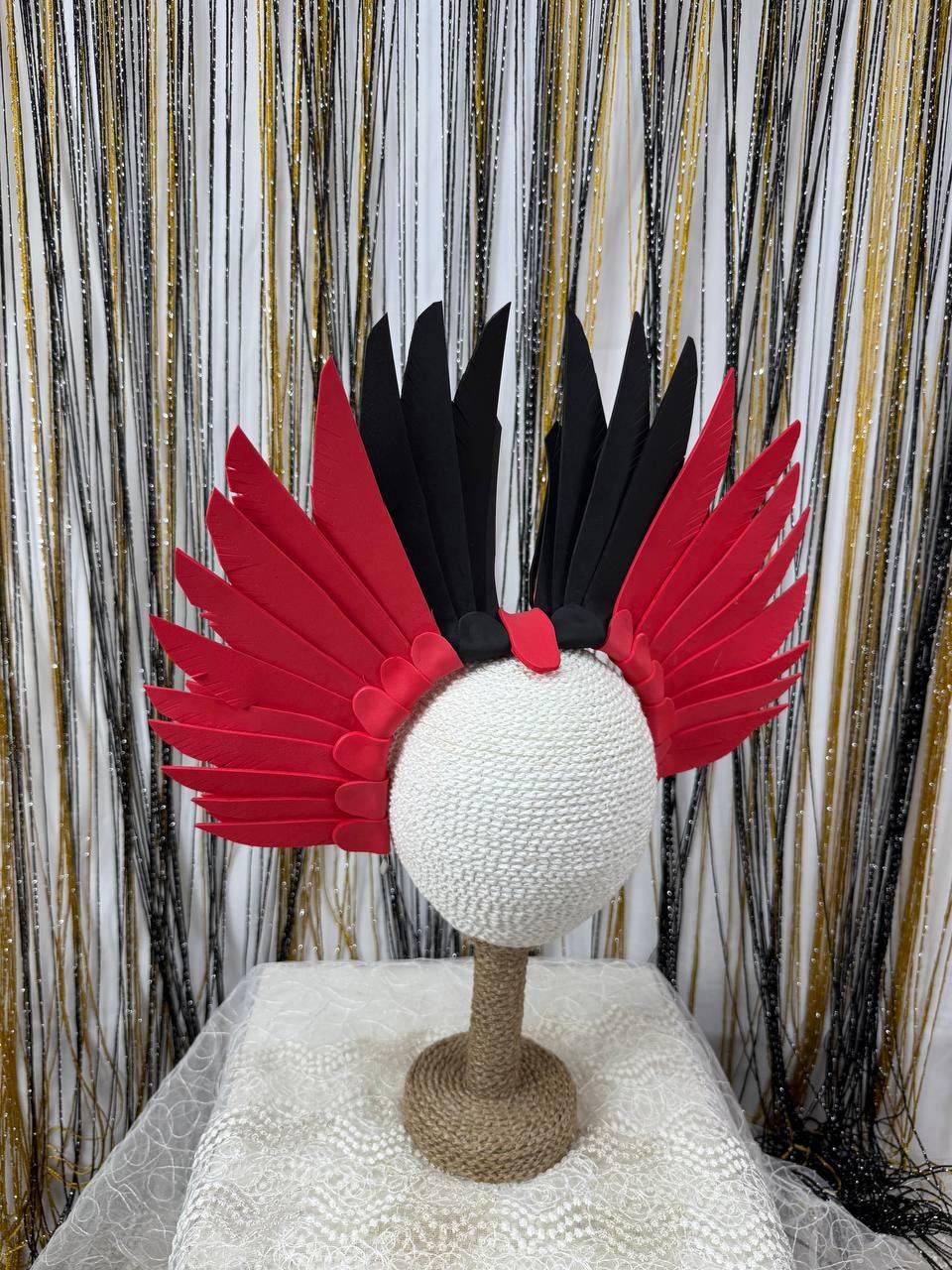 May include: A red and black feather headdress with a white sphere base. The headdress features red and black feathers arranged in a wing-like design. The base is a white sphere on a brown stand.