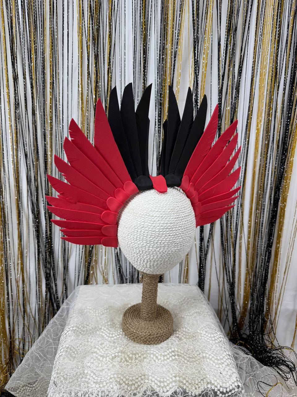 May include: A decorative headdress featuring red and black feather-like elements. The headpiece is mounted on a white sphere, supported by a jute-wrapped stand. The background includes a shimmering gold and black curtain.