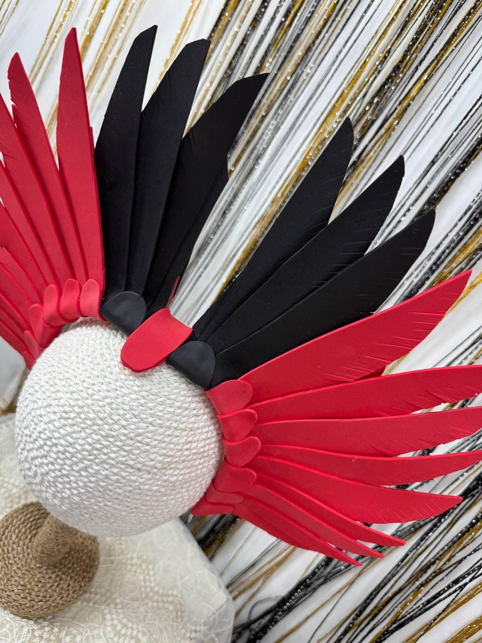 May include: A decorative headpiece featuring a white sphere base adorned with red and black feather-like elements. The feathers are arranged in a wing-like pattern, creating a striking visual contrast against the white base.