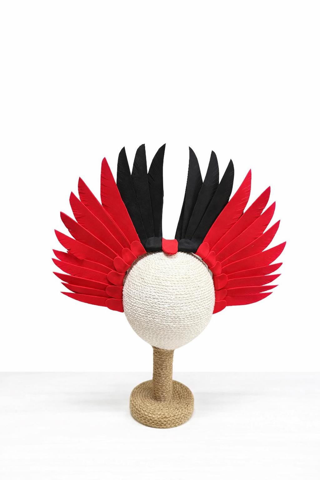 May include: A decorative headpiece with red and black feather accents. The headpiece features a white, spherical base and is displayed on a stand. The feathers are arranged in a wing-like design, creating a striking visual effect.