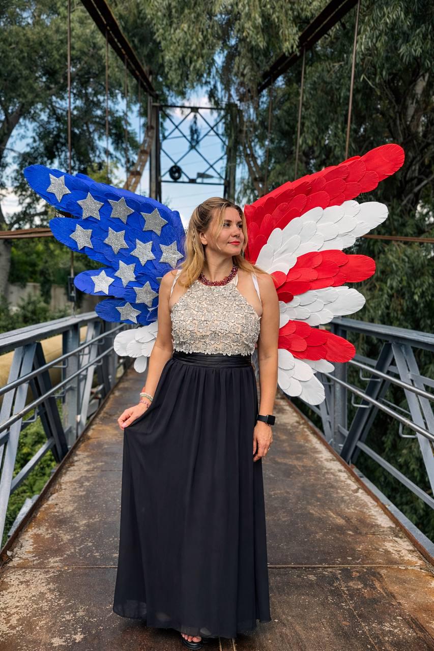 May include: A woman wearing a black maxi skirt and a beaded halter top stands on a bridge. Behind her are large, decorative wings in the colors of the American flag. The wings feature blue, white, and red sections with silver star accents.