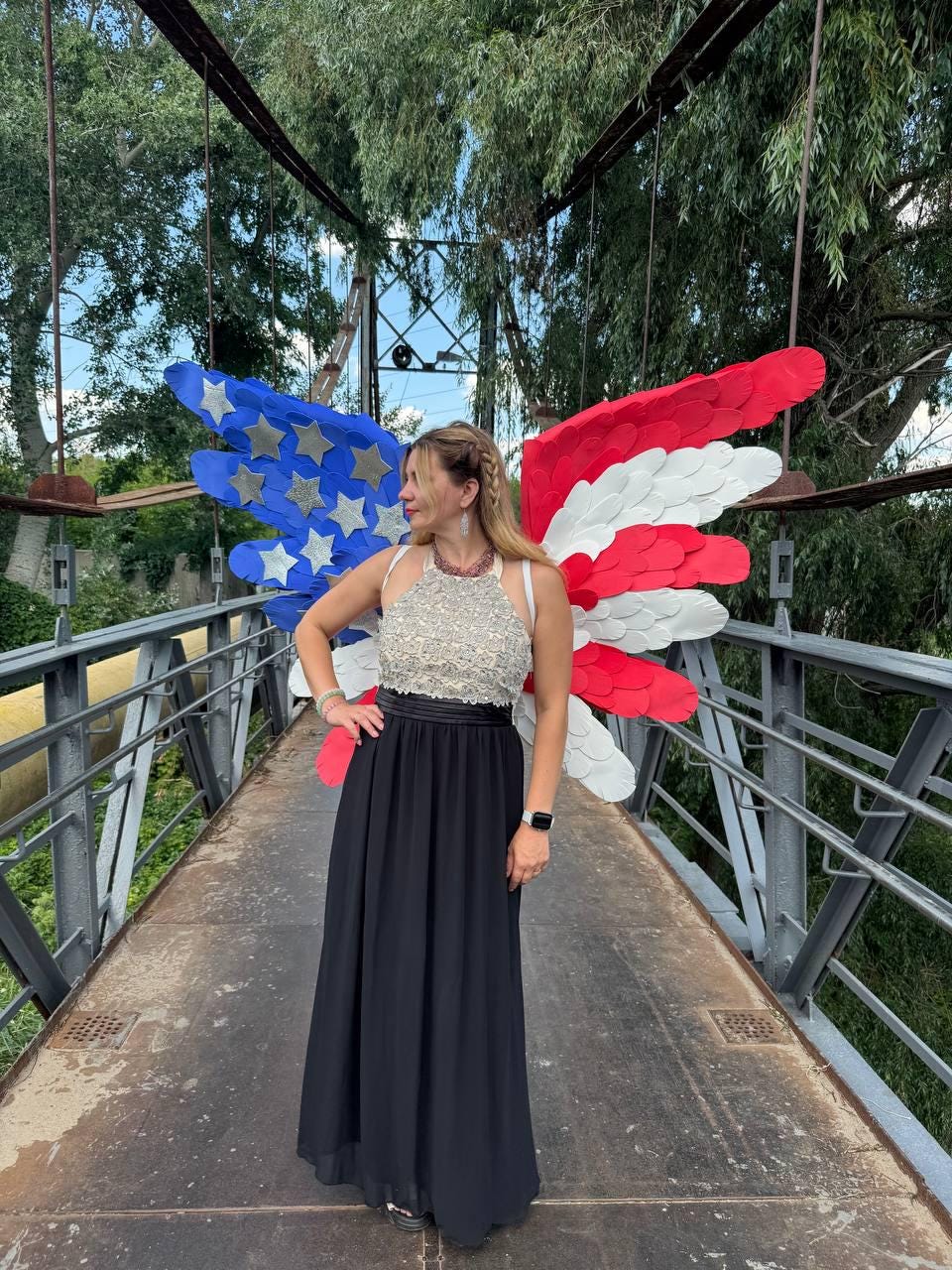 May include: A woman wearing a silver beaded halter top and a long black skirt stands on a bridge. Behind her are large, decorative wings in the style of the American flag, with blue stars and red and white stripes.