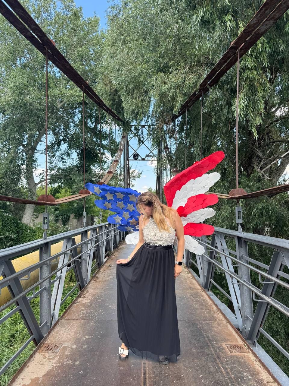 May include: A woman wearing a black maxi skirt and a white and beige top stands on a bridge. Behind her are American flag-themed wings. The bridge has metal railings and a metal walkway. The background includes trees and a blue sky.