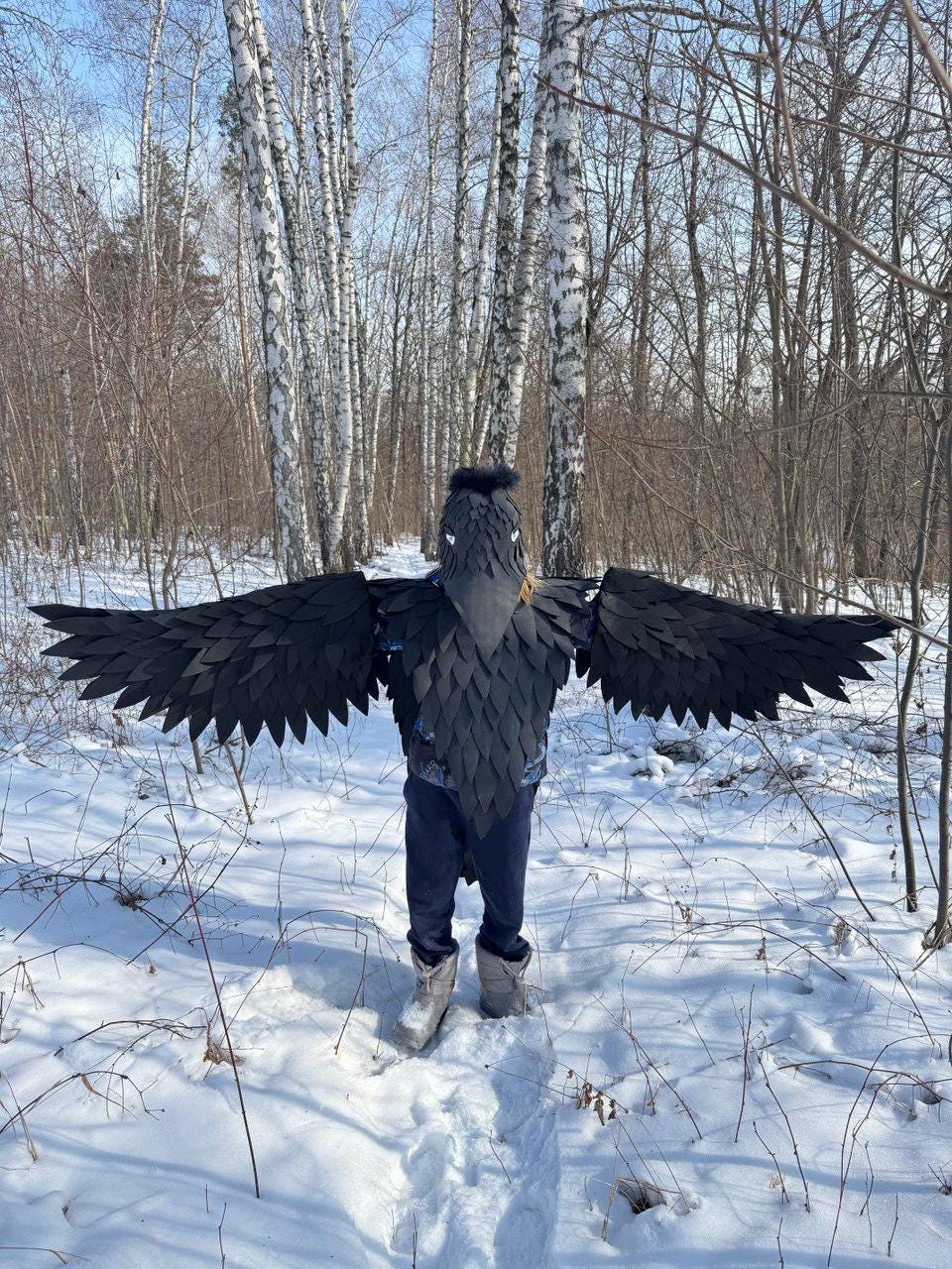May include: A person wearing a black raven costume with large, outstretched wings, standing in a snowy forest. The costume features a detailed headpiece and a textured body, creating a striking avian appearance. The wingspan is wide, and the costume is set against a backdrop of birch trees.
