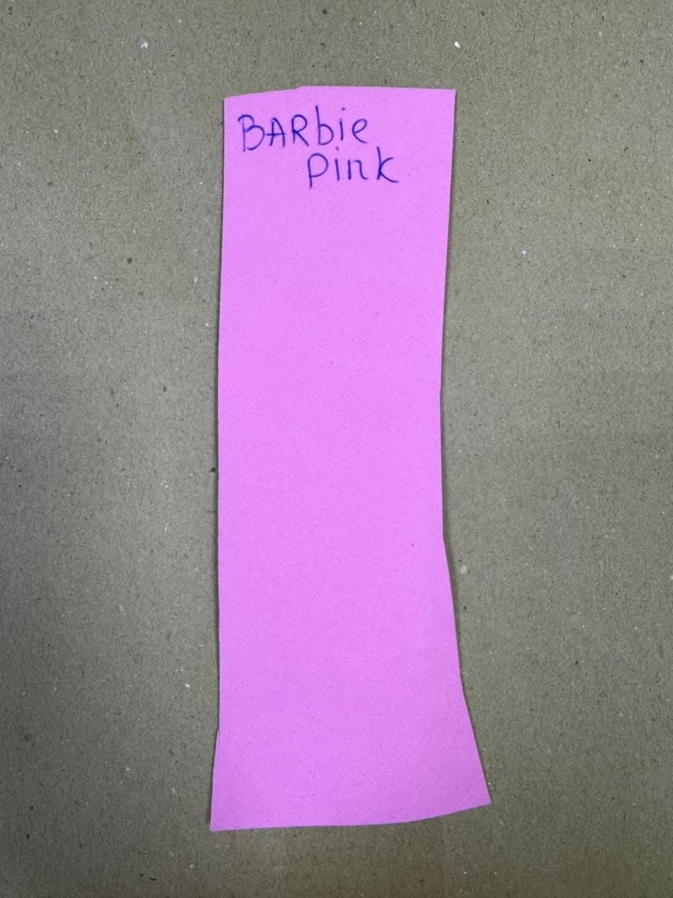 May include: A rectangular piece of Barbie pink material with the handwritten text "Barbie Pink" in dark blue ink. The material is a solid, vibrant pink color.