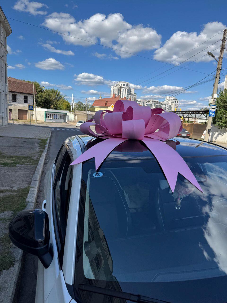 May include: A white car with a large pink bow on the roof, parked on a city street. The sky is blue with fluffy white clouds. The bow is made of multiple layers of pink ribbon.