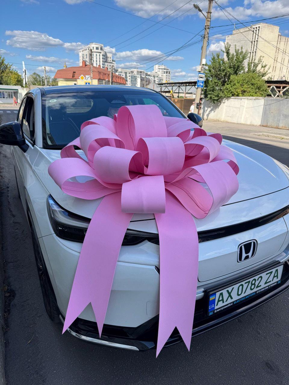 May include: A white Honda car featuring a large, decorative pink ribbon bow centered on the hood. The vehicle has a visible license plate reading AX 0782 ZA. The bow is constructed from wide, matte pink material with multiple loops and two long tails.