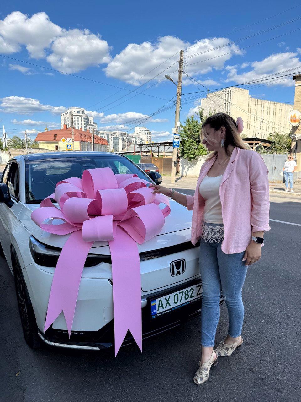 May include: A white Honda car with a large pink bow on the hood. The car is parked on a city street under a blue sky with clouds. The car has a license plate.