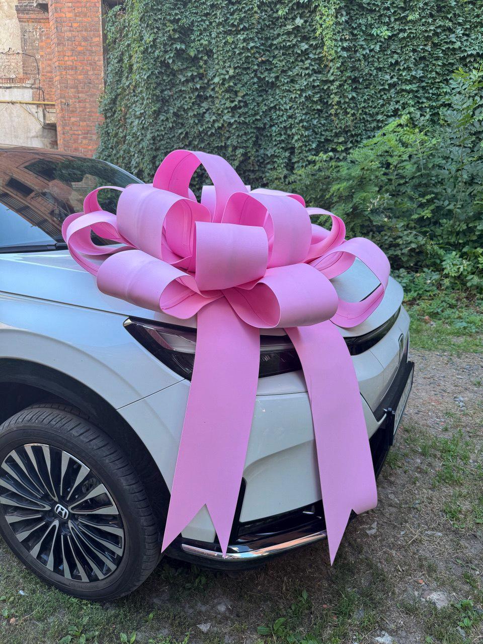 May include: A large, bright pink bow adorns the front of a white car. The bow is made of wide, looped ribbons and is positioned on the hood. The car has a black front bumper and a silver rimmed tire.