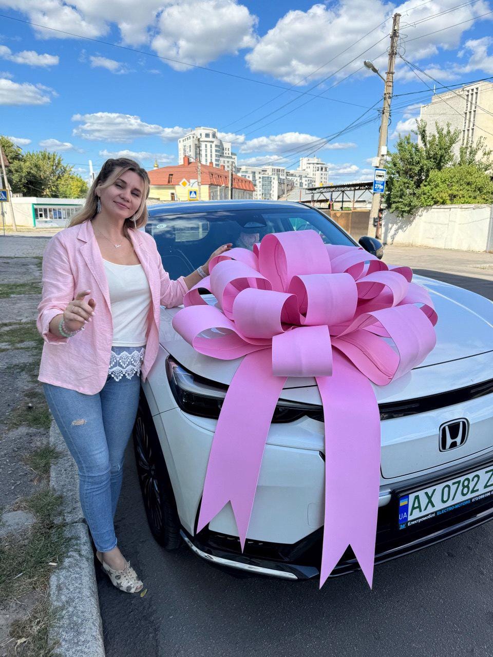 May include: A white car with a large pink bow on the hood. The car is parked on a street with a woman standing next to it. The woman is wearing a pink blazer, white top, and blue jeans. The sky is blue with clouds.