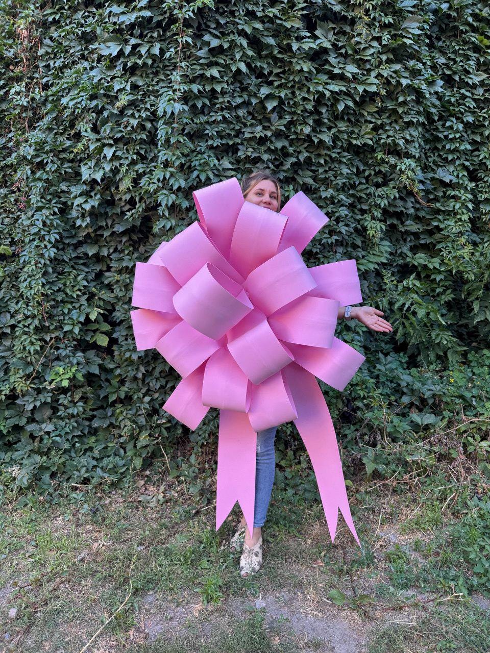 May include: A large, light pink bow with long, flowing ribbons. The bow is the focal point, with a person partially visible behind it. The background features lush green foliage and grass, creating a natural setting.