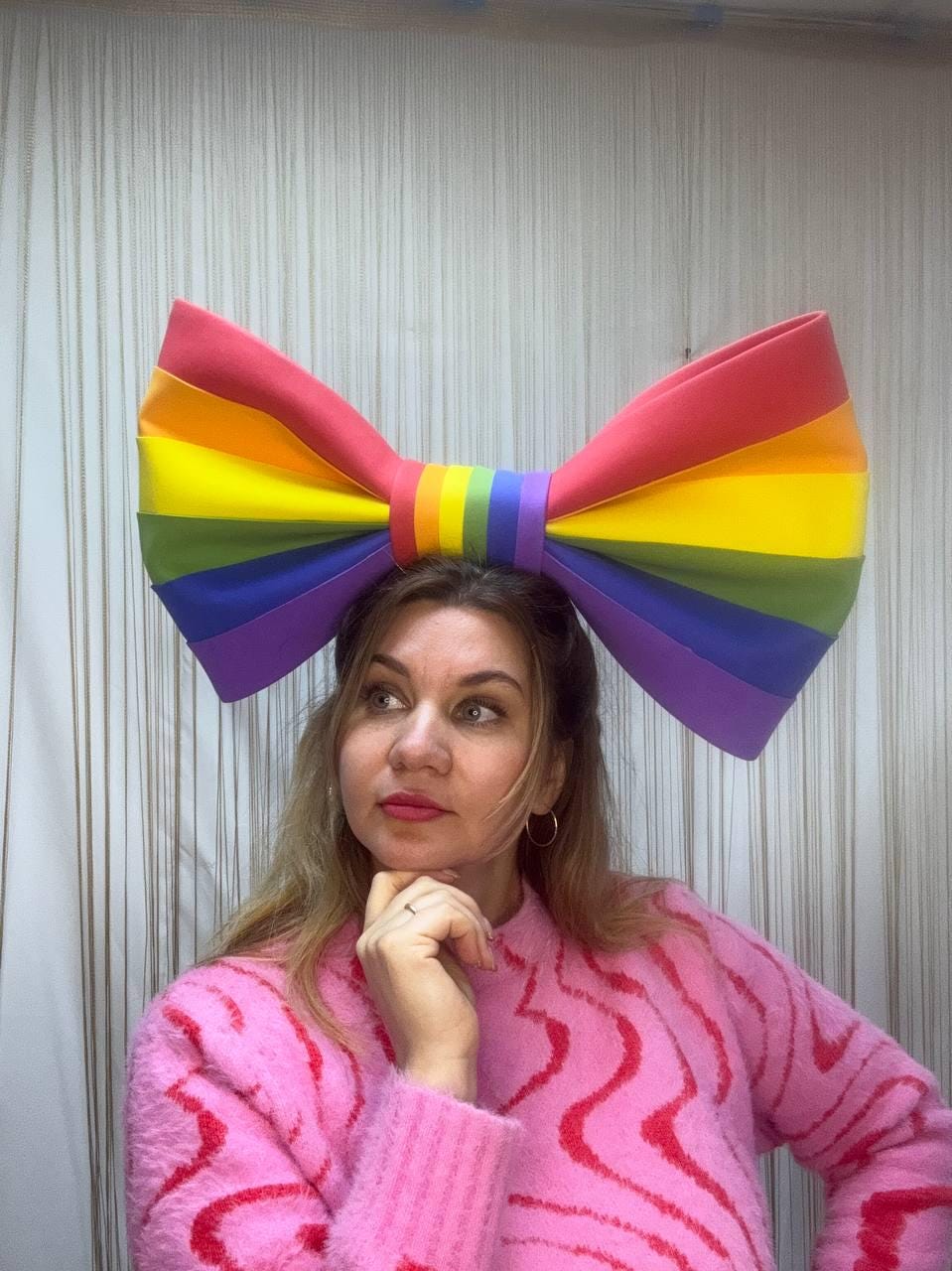 May include: A large, colorful bow in the colors of the rainbow is worn on the head. The bow is red, orange, yellow, green, blue, and purple. The person is wearing a pink sweater.