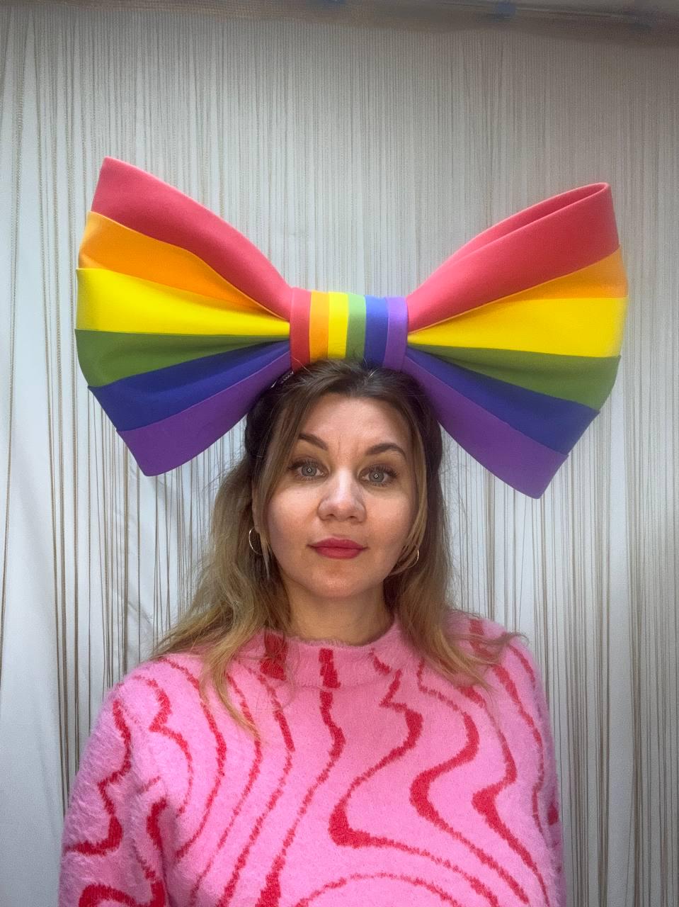May include: A large, colorful rainbow bow headband. The bow features the colors of the Pride flag: red, orange, yellow, green, blue, and purple. The bow is worn on the head. The person is wearing a pink sweater with a red wavy pattern.