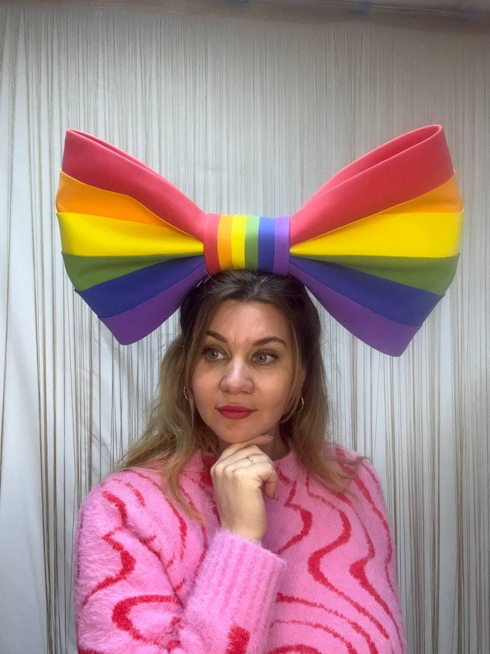 May include: A large, colorful rainbow bow, featuring red, orange, yellow, green, blue, and purple hues. The bow is worn on the head. The person is wearing a pink sweater with a red wavy pattern.
