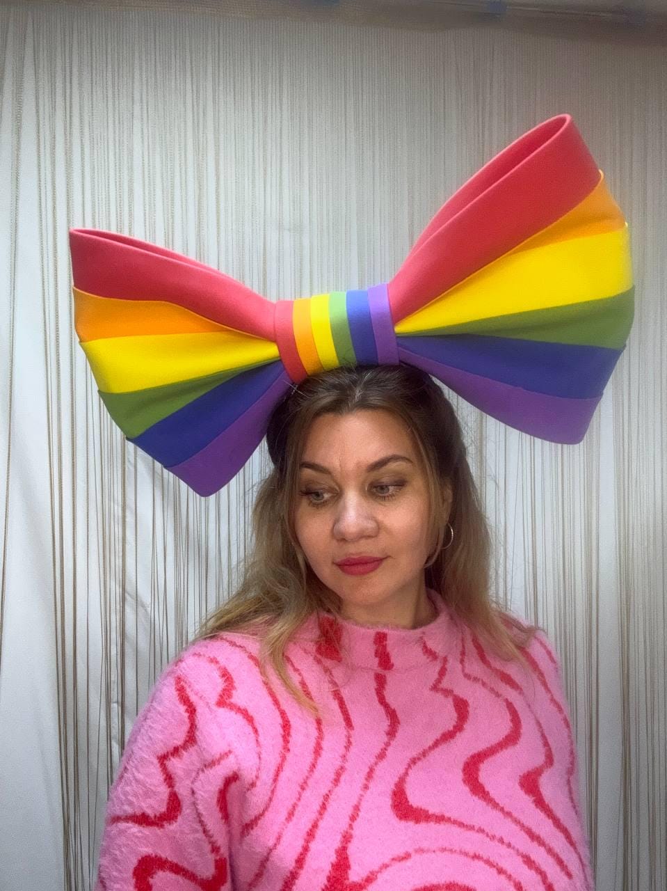 May include: A large, rainbow-colored bow headband. The bow features the colors of the rainbow: red, orange, yellow, green, blue, and purple. The bow is worn on the head. The person is wearing a pink sweater.