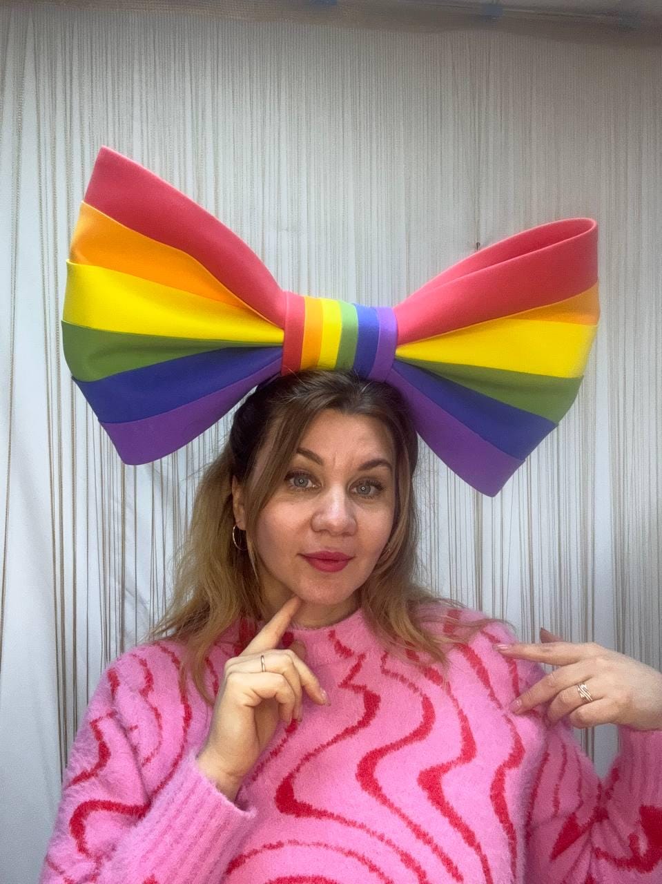 May include: A large, rainbow-colored bow, featuring red, orange, yellow, green, blue, and purple stripes. The bow is worn on the head. The person is wearing a pink sweater.