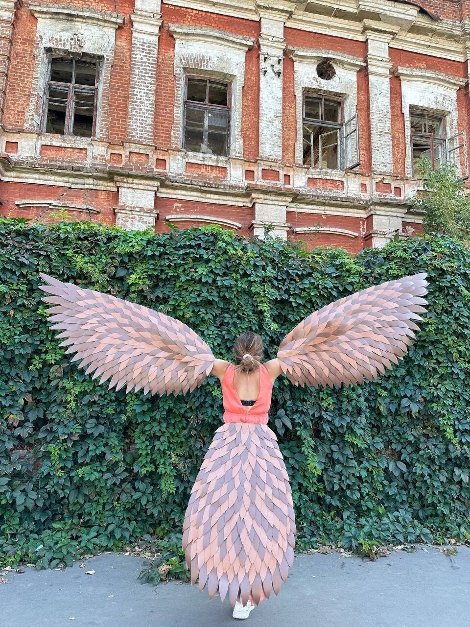 May include: A person wearing a costume with large, layered wings and a matching skirt, standing in front of a wall of green foliage. The wings and skirt are shades of brown and pink. The person is wearing a coral top.